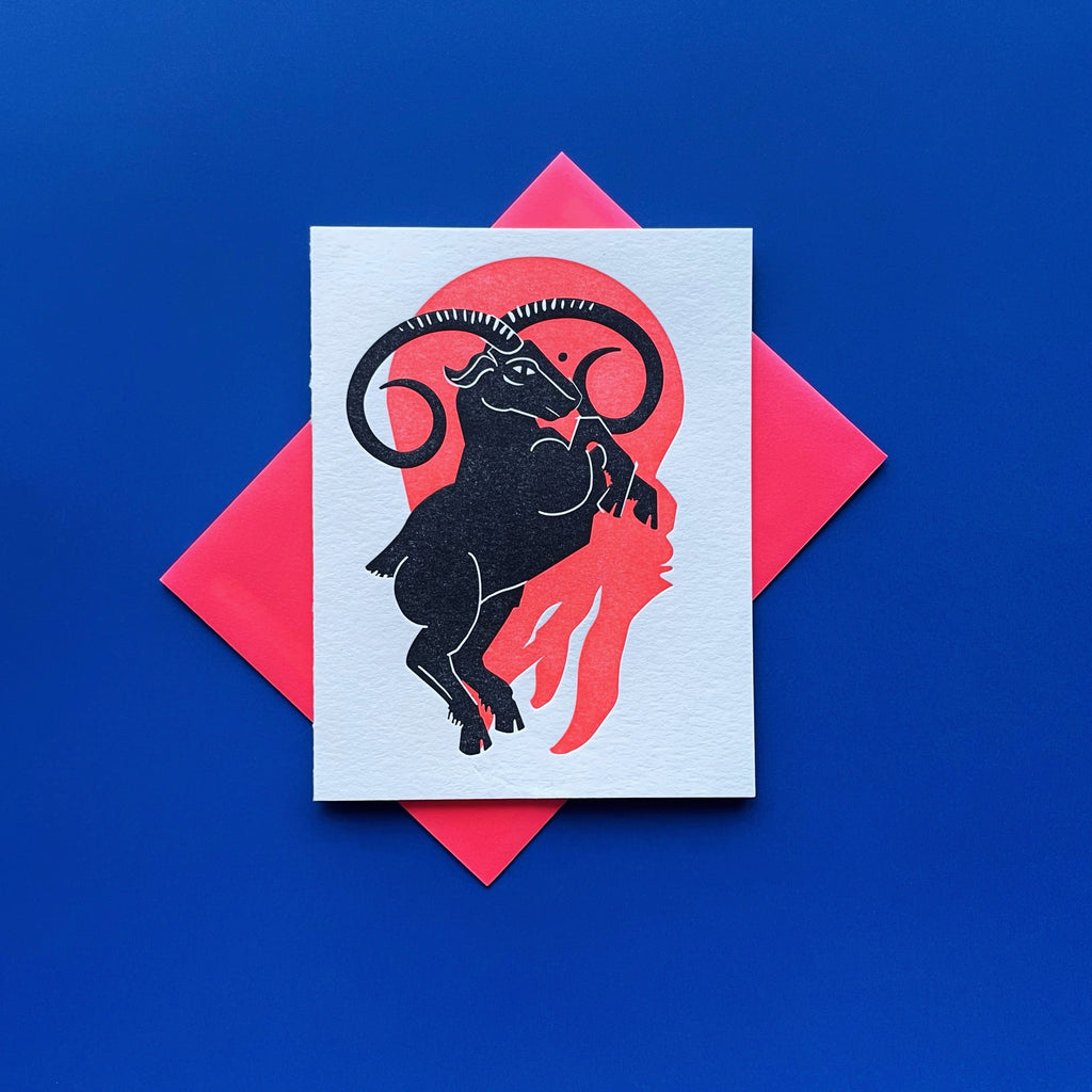 Aquarius letterpress birthday card. Black urn with all seeing eye pouring water into a esoteric waterfall. Neon red pattern in background symbolizing fire element. Shown with a red envelope on a royal blue background.