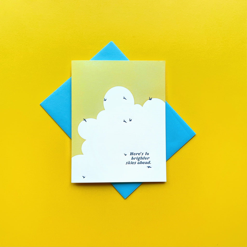 birds flying in sky with words "here's to brighter skies ahead" card featured on a yellow background shown with a neon blue envelope