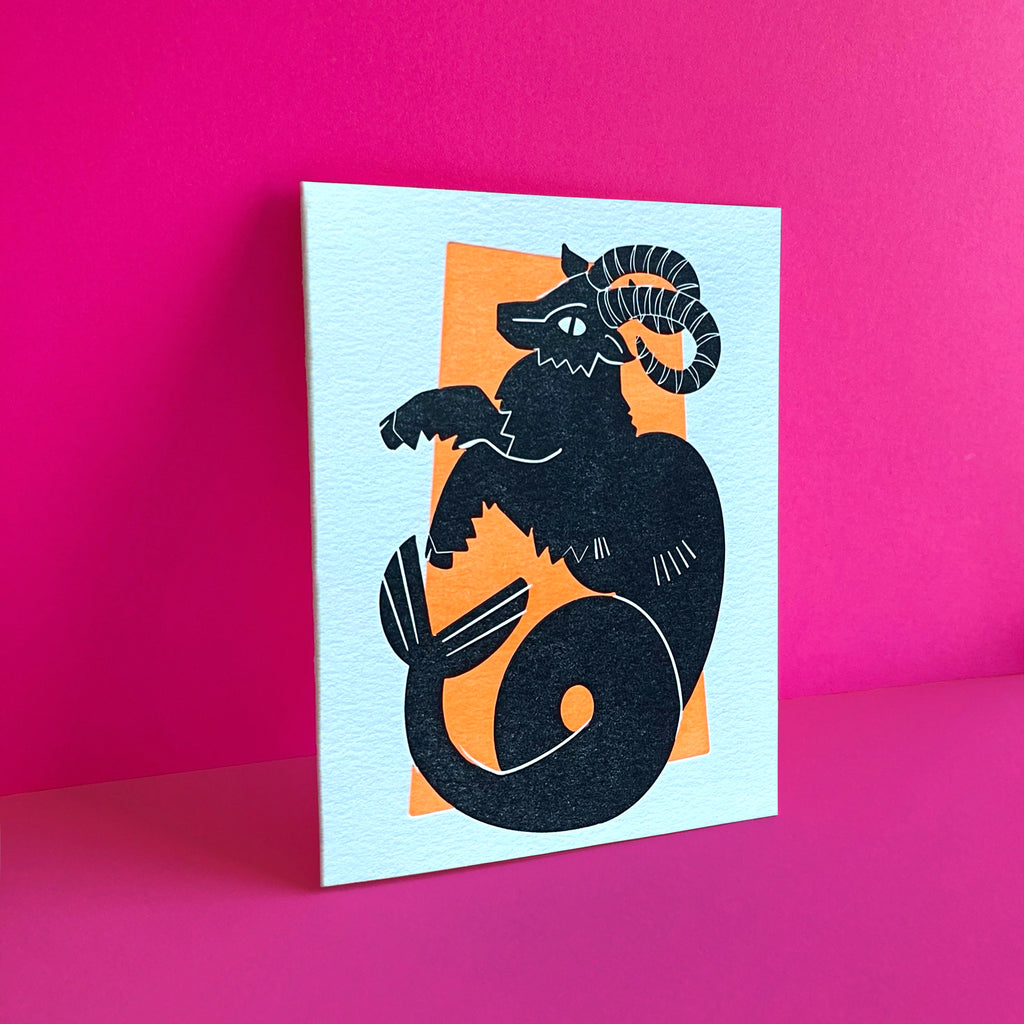 Capricorn zodiac letterpress birthday card. Black letterpress printed sea goat. Neon orange pattern in background symbolizing earth element. Shown on a bright pink background.