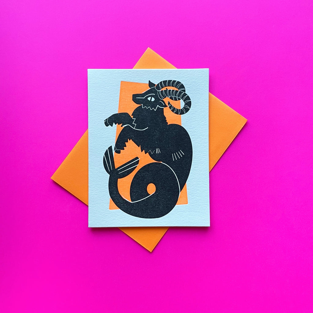 Capricorn zodiac letterpress birthday card. Black letterpress printed sea goat. Neon orange pattern in background symbolizing earth element. Shown with a neon orange envelope on a bright pink background.