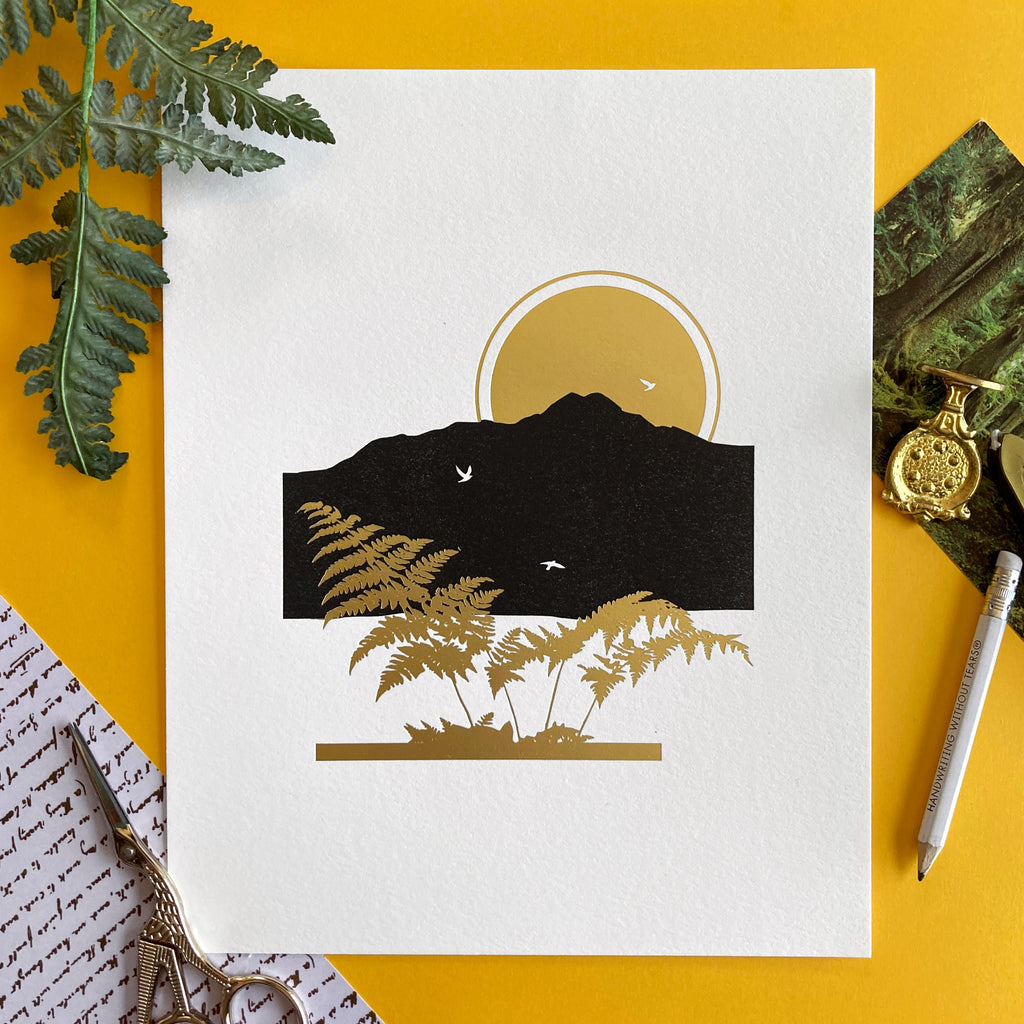 Gold foil letterpress art print of mountains and a golden fern with the sun behind the mountains. Yellow background with northwest ferns and stationery items