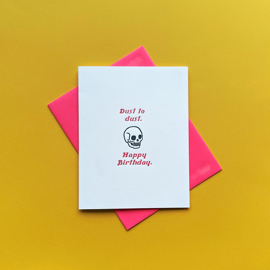 Skull with red lettering "dust to dust, happy birthday" funny letterpress birthday card shown on a yellow background with a neon red envelope