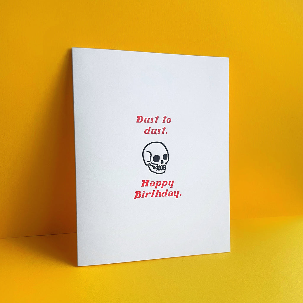 Skull with red lettering "dust to dust, happy birthday" funny letterpress birthday card shown on a yellow background