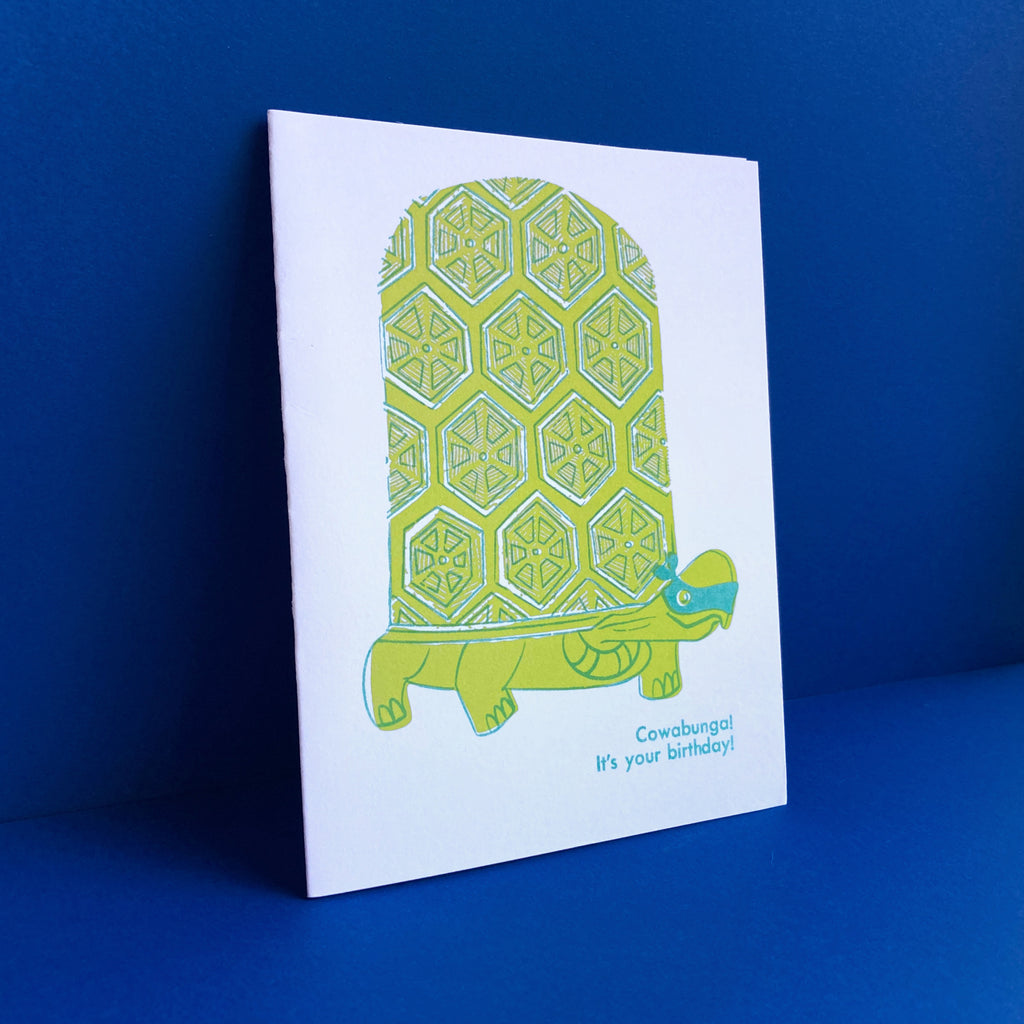 Turtle wearing TMNT style ninja face mask with the phrase "Cowabunga! It's your birthday" risograph greeting card. Shown on a royal blue background