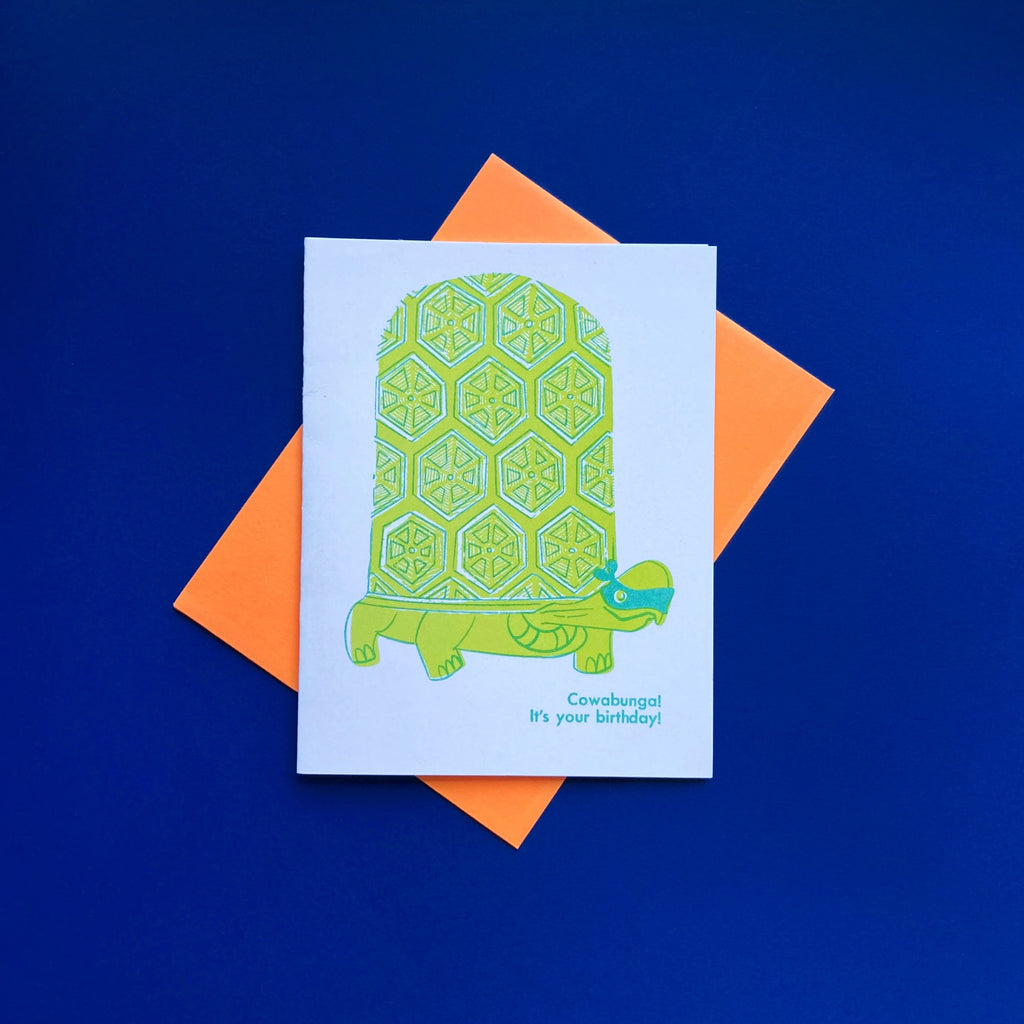 Turtle wearing TMNT style ninja face mask with the phrase "Cowabunga! It's your birthday" risograph greeting card. Shown with a neon orange envelope on a royal blue background